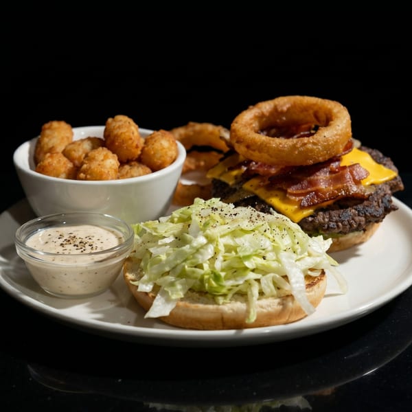 A bacon cheeseburger with onion rings, tater tots, and a side of dipping sauce on a white plate.