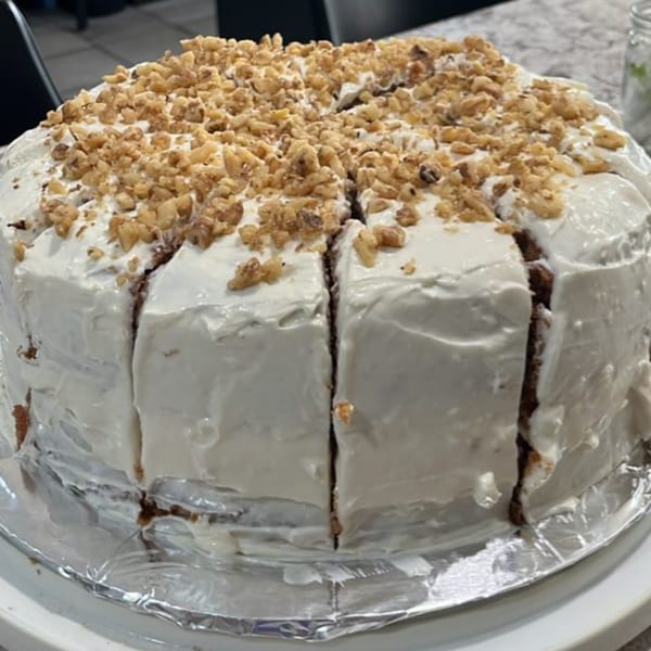 A large, round carrot cake with white frosting and chopped walnuts on top, partially sliced.